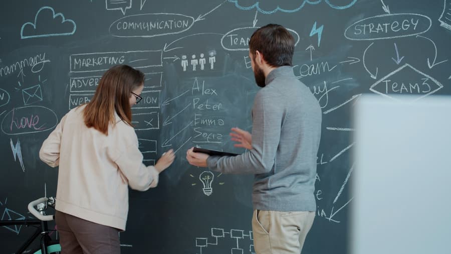 Two people stand at a chalkboard covered in diagrams, one pointing at a marketing sketch while the other holds a tablet.